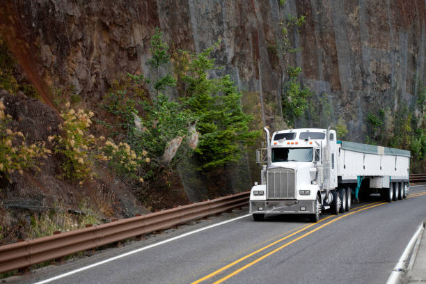 Big rig white classic American model bonnet semi truck tractor with plastic covered long bulk semi trailer driving on the mountain road with rock mountain cliff wall in Columbia River Gorge area